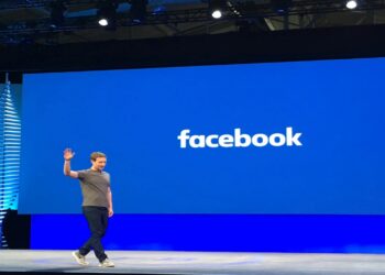 Mark Zuckerberg acenando em um evento do Facebook com um grande logotipo ao fundo.