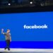 Mark Zuckerberg acenando em um evento do Facebook com um grande logotipo ao fundo.