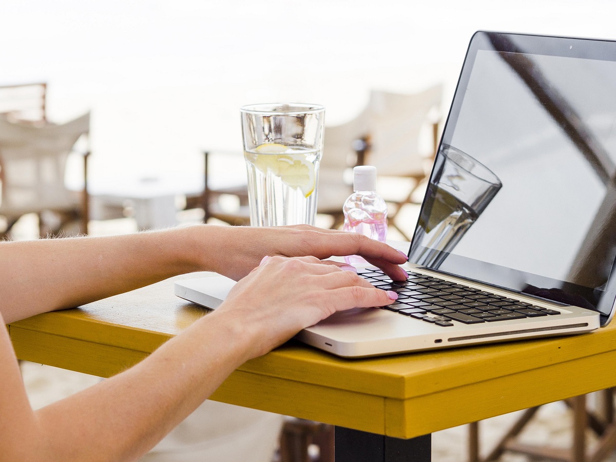 Pessoa digitando no laptop por dicas de sites em uma mesa ao ar livre, com uma bebida e um frasco de água ao lado.