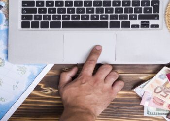 Imagem de uma mão tocando o touchpad de um laptop, com um mapa, câmera, dinheiro e passaporte ao redor dos sites.