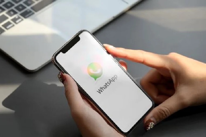 Pessoa segurando um celular com o logo do WhatsApp visível na tela, com um laptop ao fundo, representando o uso do app em fraudes.