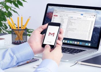 Pessoa acessando e organizando e-mails no Gmail usando smartphone e laptop em ambiente de trabalho.